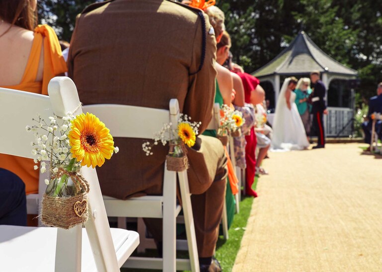 A yellow flower in a little jam jar saying love on it with the bride and groom in the background during their outdoor wedding ceremony at Hungarian Hall on a Wedding Day photographed by Suffolk Wedding Photographers Hayley Denston Photography