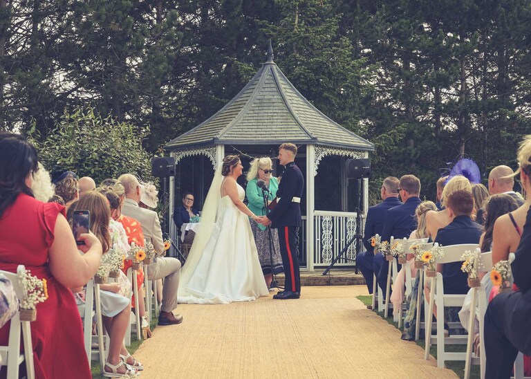 A bride and groom holding hands looking at each other during an outdoor wedding ceremony with a pergola behind them at Hungarian Hall on a Wedding Day photographed by Suffolk Wedding Photographers Hayley Denston Photography
