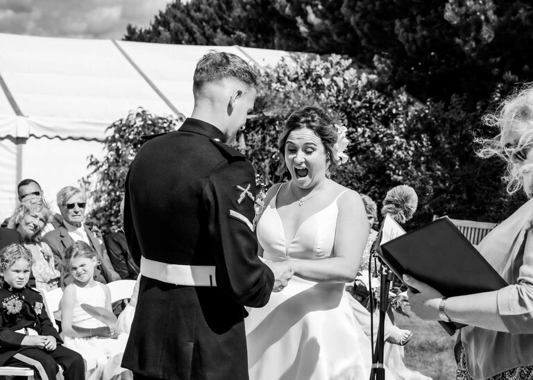 A bride gasping as she looks at her wedding ring during an outdoor ceremony at Hungarian Hall on a Wedding Day photographed by Suffolk Wedding Photographers Hayley Denston Photography