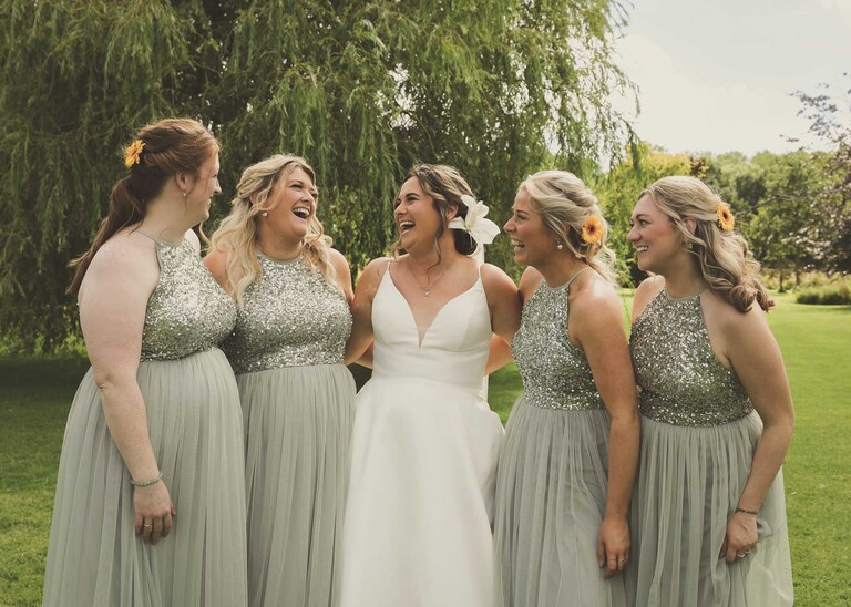 A bride laughing with her bridesmaids dressed in sage green and sparkly sequins at Hungarian Hall on a Wedding Day photographed by Suffolk Wedding Photographers Hayley Denston Photography