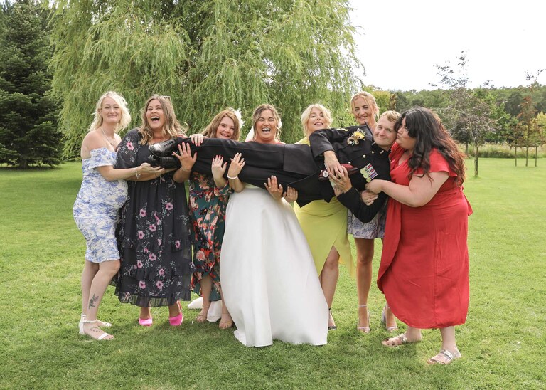A group of military wives holding the groom in a lyaing position at Hungarian Hall on a Wedding Day photographed by Suffolk Wedding Photographers Hayley Denston Photography