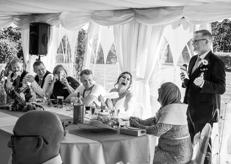 A father of the bride giving his speech whilst the bride and mother of the bride are laughing at Hungarian Hall on a Wedding Day photographed by Suffolk Wedding Photographers Hayley Denston Photography
