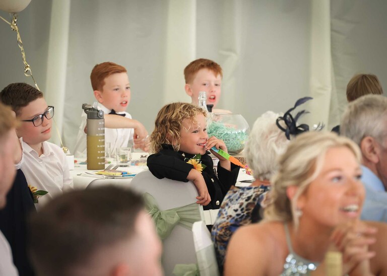 A pageboy watching his dad from a childs table giving a speech at Hungarian Hall on a Wedding Day photographed by Suffolk Wedding Photographers Hayley Denston Photography