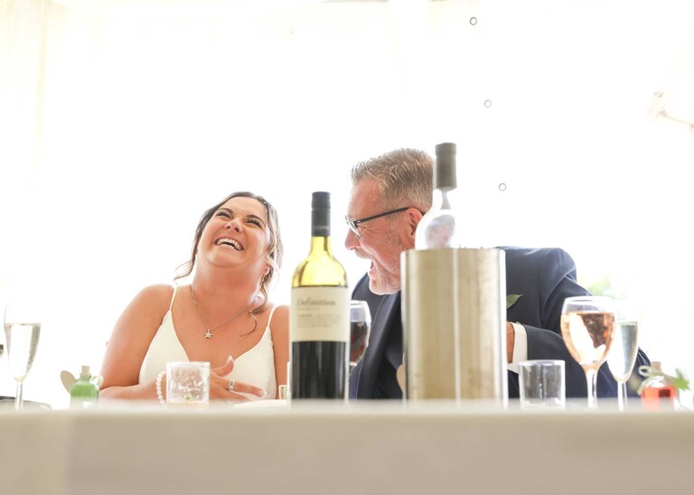 A bride and her dad laughing during the grooms speech at Hungarian Hall on a Wedding Day photographed by Suffolk Wedding Photographers Hayley Denston Photography