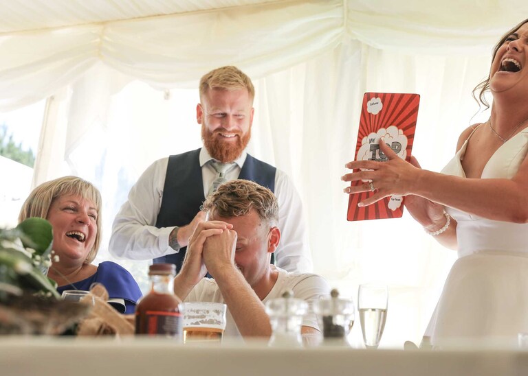 A bride and bestman laughing whilst the groom holds his head during the bestmans speech at Hungarian Hall on a Wedding Day photographed by Suffolk Wedding Photographers Hayley Denston Photography