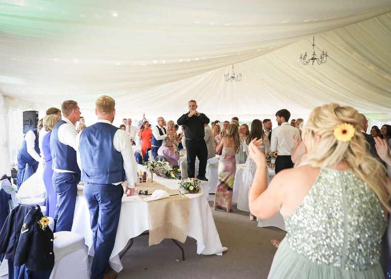 A singing waiter performing to guests whilst standing on a chair in a marquee at Hungarian Hall on a Wedding Day photographed by Suffolk Wedding Photographers Hayley Denston Photography