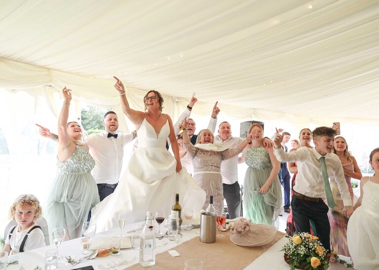 A bride stood on a chair pointing and dancing with guests during a singing waiters rendition of sex on fire at Hungarian Hall on a Wedding Day photographed by Suffolk Wedding Photographers Hayley Denston Photography