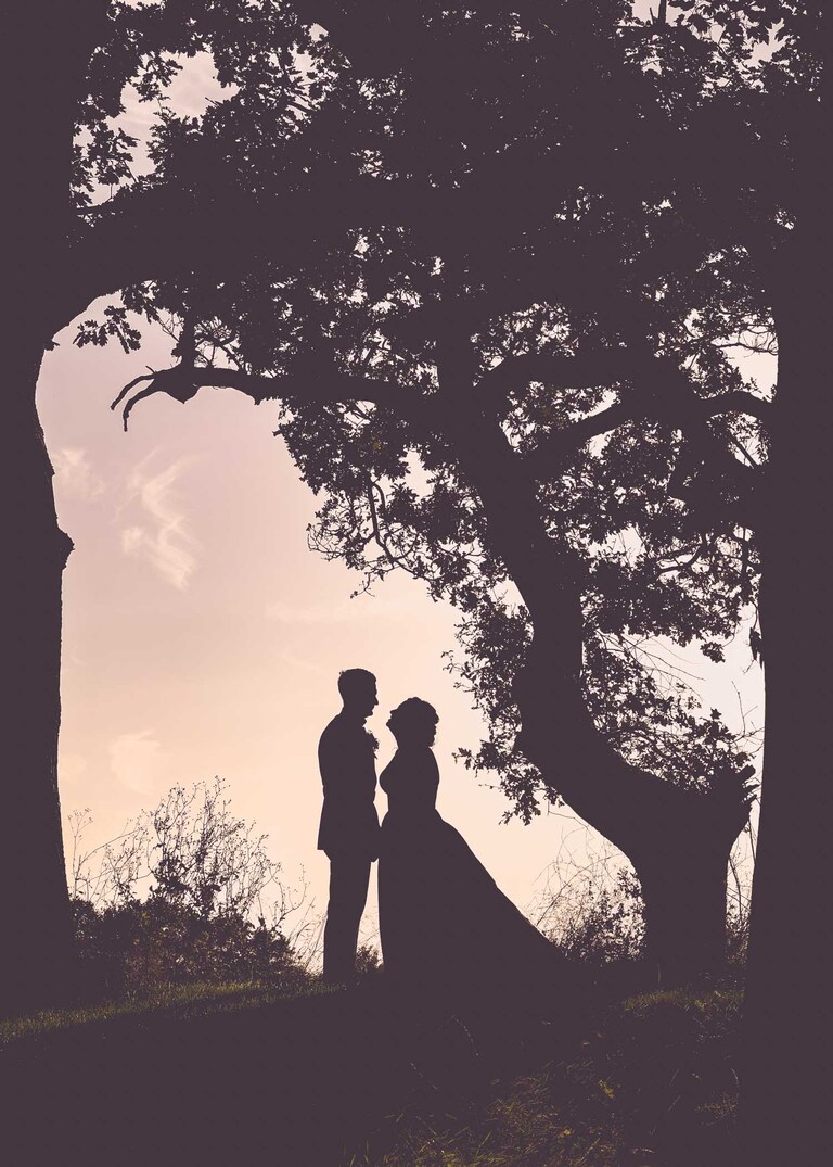 A bride and groom in silhouette stood under trees facing each other at sunset at Hungarian Hall on a Wedding Day photographed by Suffolk Wedding Photographers Hayley Denston Photography