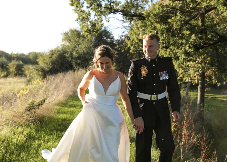 A bride and groom walking hand in hand with beautiful sunlight behind them at sunset at Hungarian Hall on a Wedding Day photographed by Suffolk Wedding Photographers Hayley Denston Photography