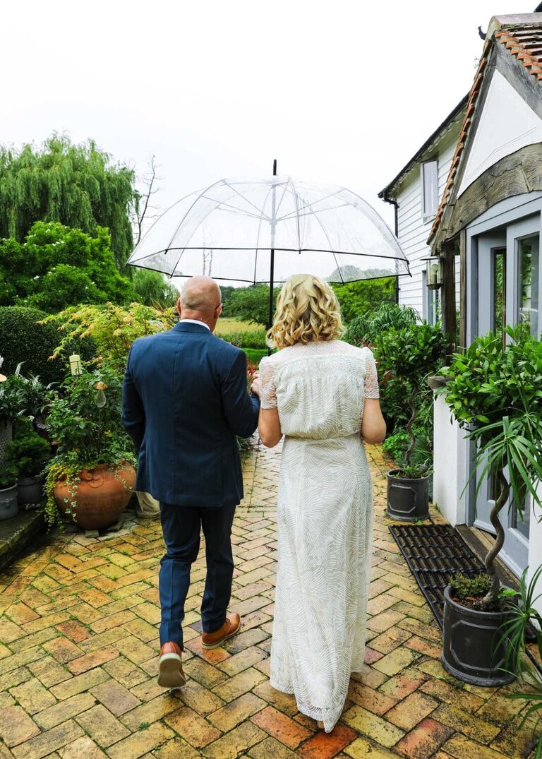 A bride and groom walking away under a clear umbrella on a rainy wedding day at Letheringham Water Mill by Suffolk Wedding Photographers Hayley Denston Photography