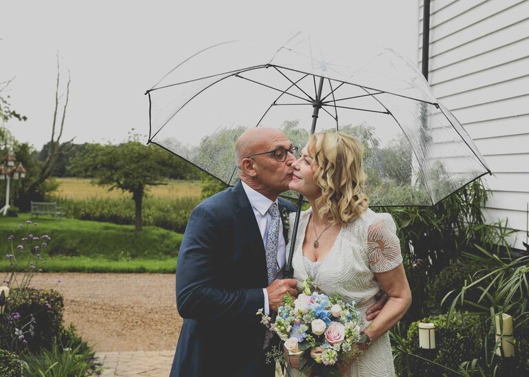 A bride and groom with the groom kissing the bride on the cheek under a clear umbrella on a rainy wedding day at Letheringham Water Mill by Suffolk Wedding Photographers Hayley Denston Photography