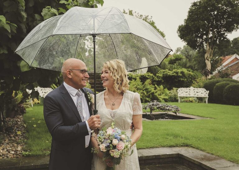 A bride and groom under a clear umbrella on a rainy wedding day at Letheringham Water Mill by Suffolk Wedding Photographers Hayley Denston Photography
