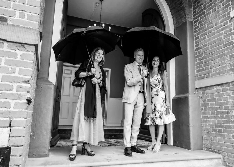 Guests stood under umbrellas before a wedding ceremony at Woodbridge Shire Hall on a rainy day photographed by Suffolk Wedding Photographers Hayley Denston Photography
