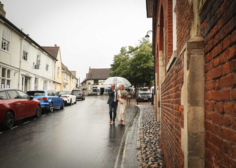 A bride and groom walking under an umbrella ready to come into their wedding ceremony at Woodbridge Shire Hall on a rainy day photographed by Suffolk Wedding Photographers Hayley Denston Photography