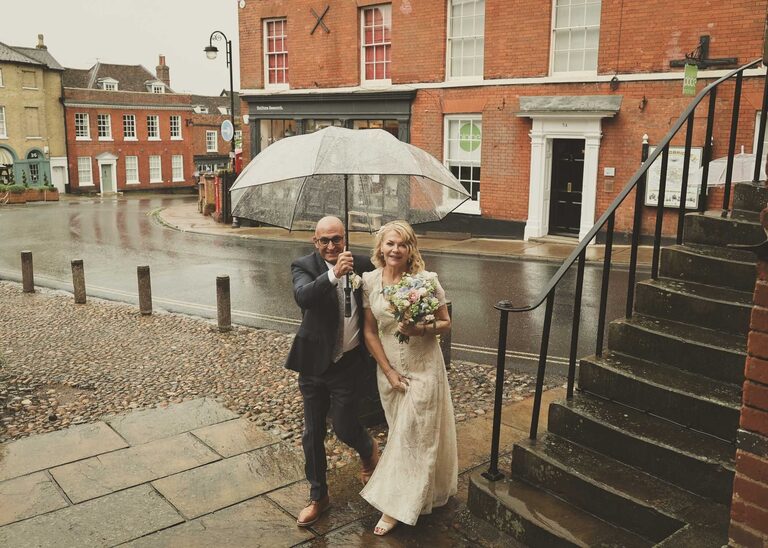 A bride and groom walking under an umbrella ready to come into their wedding ceremony at Woodbridge Shire Hall on a rainy day photographed by Suffolk Wedding Photographers Hayley Denston Photography