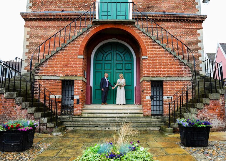 A bride and groom stood holding hands looking at each other in front of the large green doors on a rainy wedding day at Woodbridge Shire Hall photographed by Suffolk Wedding Photographers Hayley Denston Photography