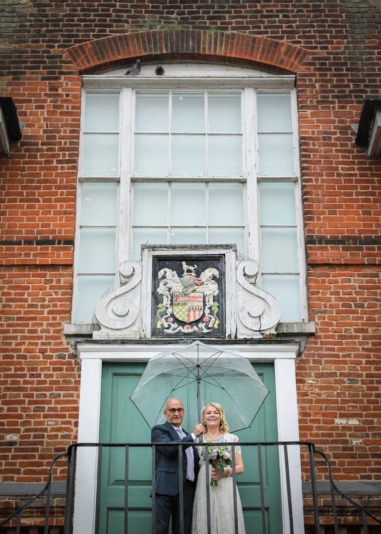 A bride and groom stood at the top of the Shire Hall under a clear umbrella on a rainy wedding day at Woodbridge Shire Hall photographed by Suffolk Wedding Photographers Hayley Denston Photography