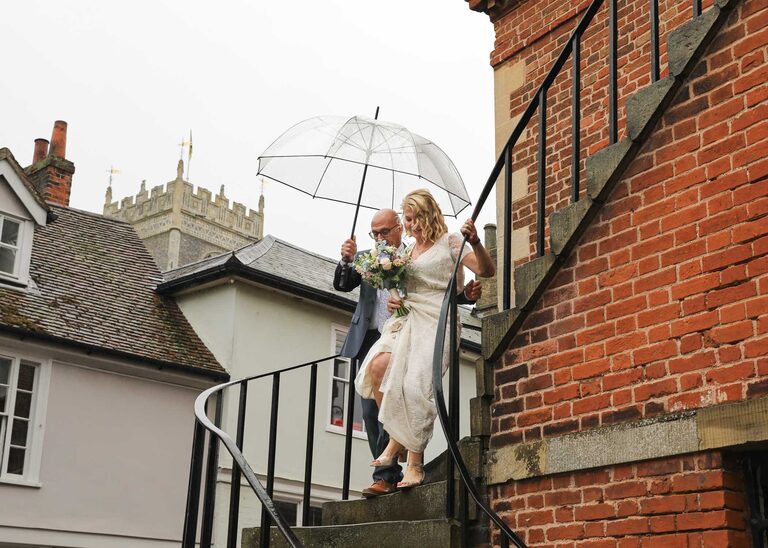 A bride and groom walking down the steps on a rainy wedding day at Woodbridge Shire Hall photographed by Suffolk Wedding Photographers Hayley Denston Photography