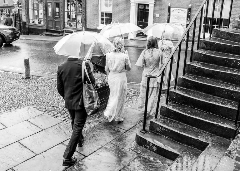 A bridal party walking under clear umbrellas on a rainy wedding day at Woodbridge Shire Hall photographed by Suffolk Wedding Photographers Hayley Denston Photography