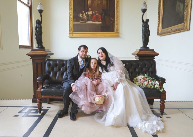 A bride and groom and their daughter the flowergirl sat ona leather sofa at Ipswich Town Hall taken by Suffolk Wedding Photographer Hayley Denston Photography