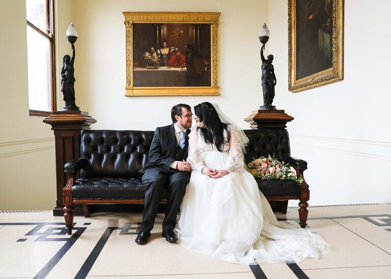 A bride and groom looking at each other sat on a leather sofa on their wedding day at Ipswich Town Hall taken by Suffolk Wedding Photographer Hayley Denston Photography
