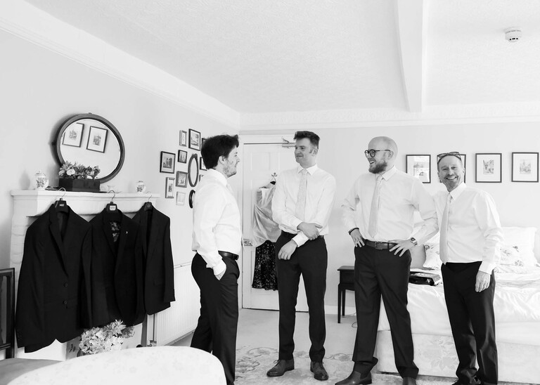 A black and white photo of a groom and groomsmen getting ready in a bedroom on his wedding day at Bressingham Hall and High Barn captured by Suffolk Wedding Photographers Hayley Denston Photography