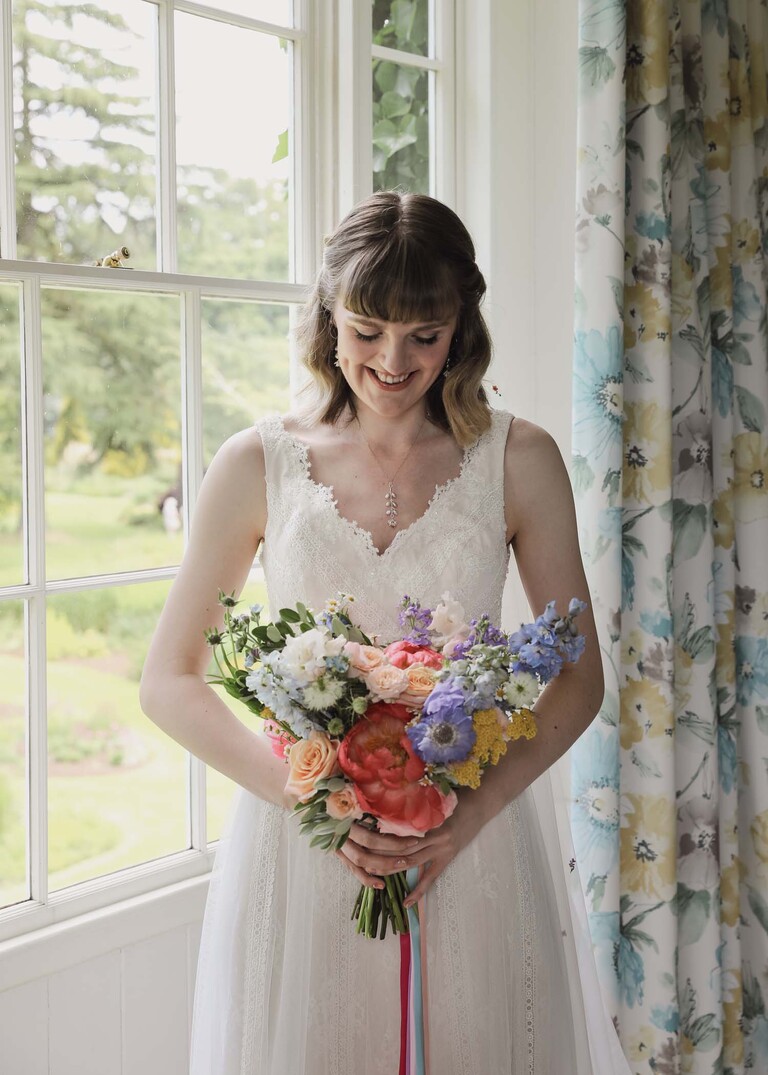 A bride stood in front of a sash window on her wedding day looking down at her rainbow coloured floral bouquet on a wedding day at Bressingham Hall and High Barn captured by Suffolk Wedding Photographers Hayley Denston Photography