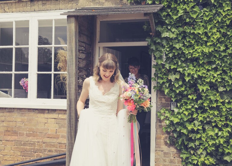 A bride walking over to her wedding ceremony with a bright rainbow coloured floral bouquet on a wedding day at Bressingham Hall and High Barn captured by Suffolk Wedding Photographers Hayley Denston Photography