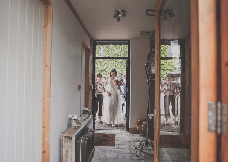 A bride walking over to her wedding ceremony with a bright rainbow coloured floral bouquet on a wedding day at Bressingham Hall and High Barn captured by Suffolk Wedding Photographers Hayley Denston Photography