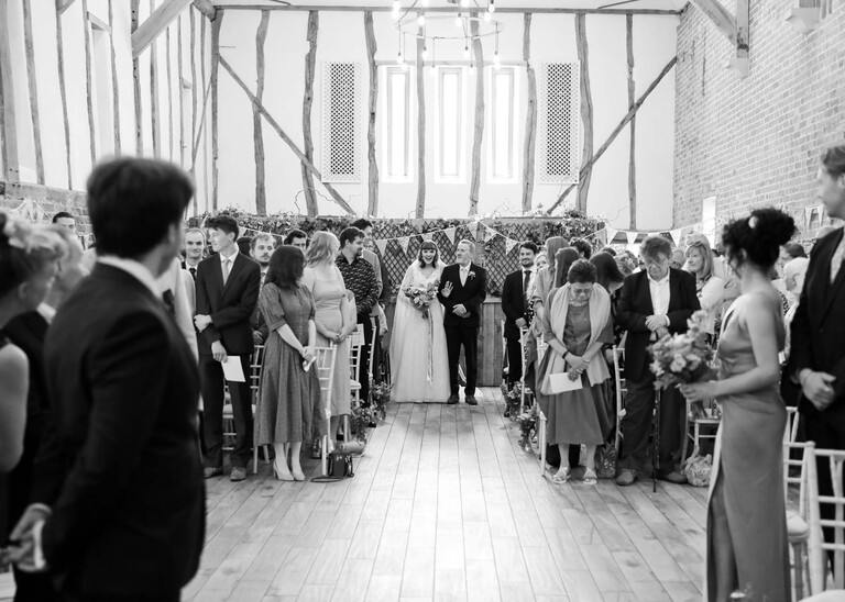 A black and white photo of a bride waving at her groom from the end of the aisle on a wedding day at Bressingham Hall and High Barn captured by Suffolk Wedding Photographers Hayley Denston Photography