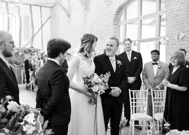 A black and white photo of a bride looking at her dad at the end of the aisle once she has reached the groom on a wedding day at Bressingham Hall and High Barn captured by Suffolk Wedding Photographers Hayley Denston Photography