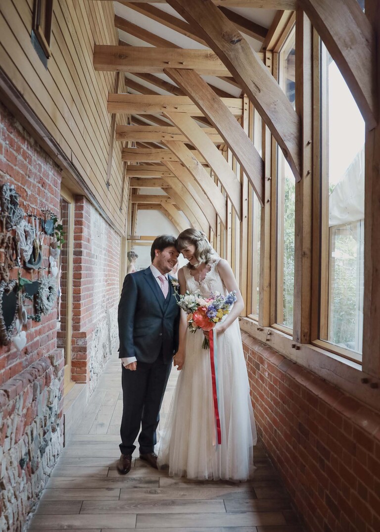 A bride and groom stood in a beautiful vaulted wooden corridor with their heads touching each other on a wedding day at Bressingham Hall and High Barn captured by Suffolk Wedding Photographers Hayley Denston Photography