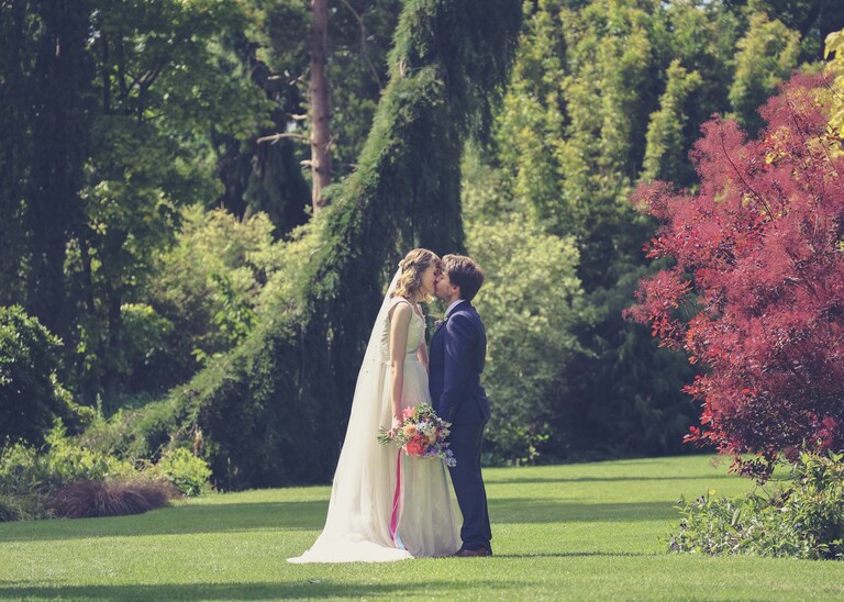 A bride and groom stood in gardens kissing with bright floral bouquet on a wedding day at Bressingham Hall and High Barn captured by Suffolk Wedding Photographers Hayley Denston Photography