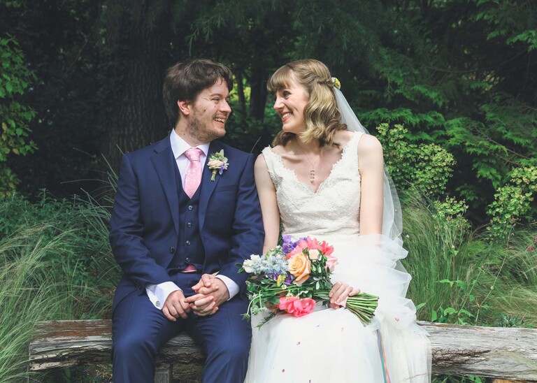 A bride and groom sat on a bench in Bressingham gardens looking at each other on a wedding day at Bressingham Hall and High Barn captured by Suffolk Wedding Photographers Hayley Denston Photography
