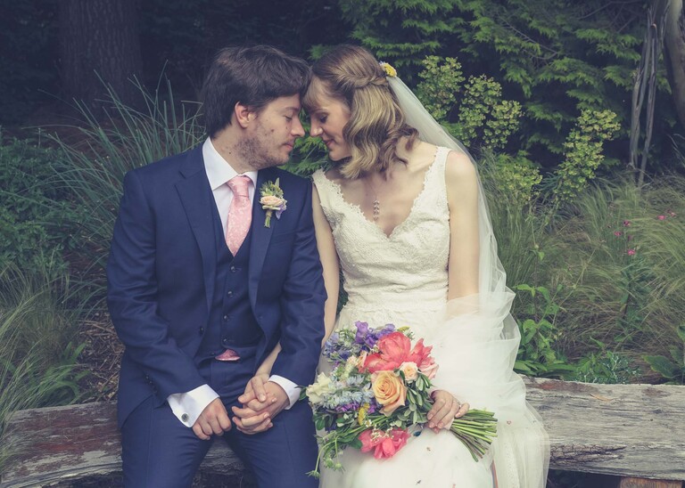 A bride and groom sat on a bench in Bressingham gardens with their heads touching each other on a wedding day at Bressingham Hall and High Barn captured by Suffolk Wedding Photographers Hayley Denston Photography