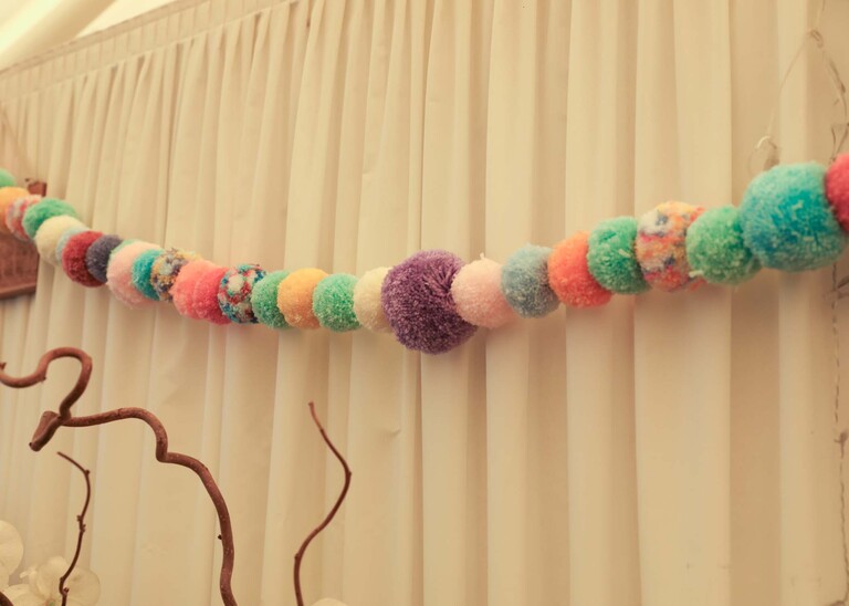 A multicoloured pompom garland in a wedding marquee on a wedding day at Bressingham Hall and High Barn captured by Suffolk Wedding Photographers Hayley Denston Photography