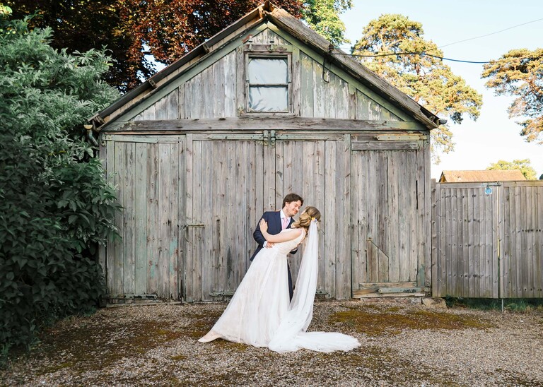 A groom and bride rehearsing their first dance and a bridal dip in front of a rustic old wooden shed in Bressingham Gardens during golden hour on a wedding day at Bressingham Hall and High Barn captured by Suffolk Wedding Photographers Hayley Denston Photography
