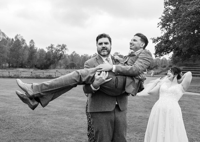 A black and white photo of a bestman carrying a groom in his arms on a wedding day at Easton Grange captured by Suffolk Wedding Photographers Hayley Denston Photography
