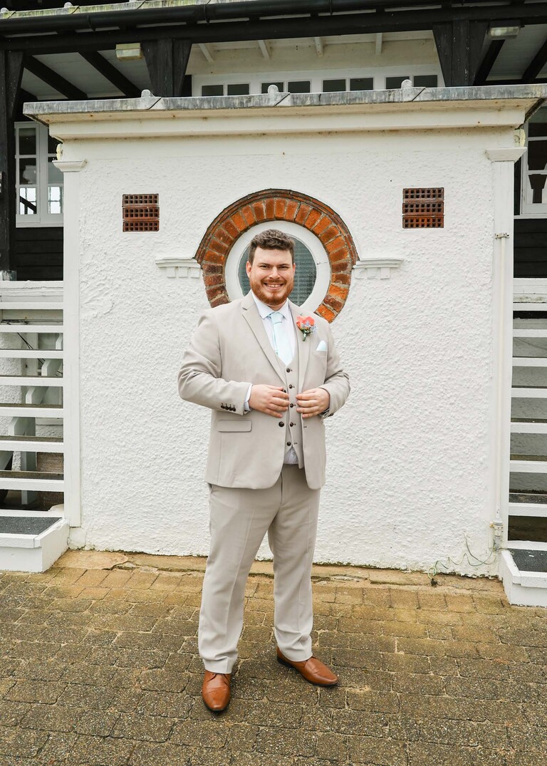 A groom stood in a pale beige suit outside his wedding venue on his wedding day at Thorpeness Country Club captured by Suffolk Wedding Photographers Hayley Denston Photography
