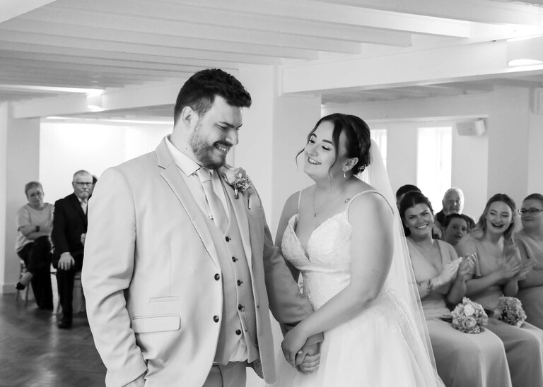 A black and white photo of the bride looking at her groom just after they have shared their first kiss curing the wedding ceremony at their wedding day at Thorpeness Country Club captured by Suffolk Wedding Photographers Hayley Denston Photography