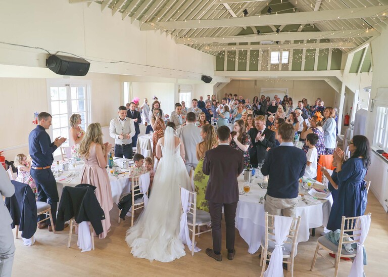 A bride and groom walking through their guests at their wedding breakfast to find their seats on a wedding day at Thorpeness Country Club captured by Suffolk Wedding Photographers Hayley Denston Photography