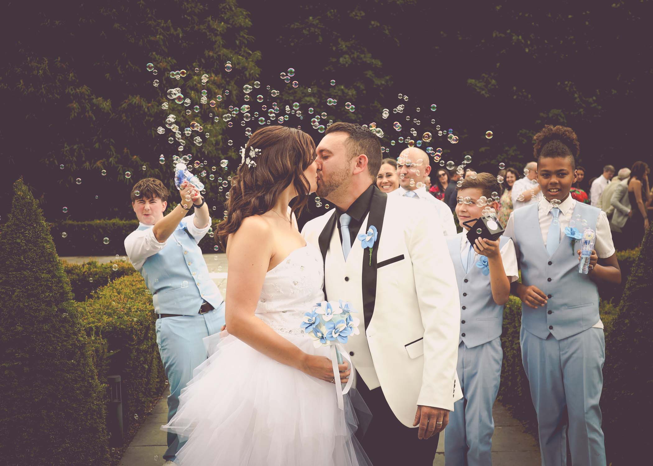 A bride and groom kissing with bubbles surrounding them after walking down the aisle during their outdoor wedding ceremony on a wedding day at Swynford Manor in Suffolk Captured by Suffolk Wedding Photographer Hayley Denston Photography