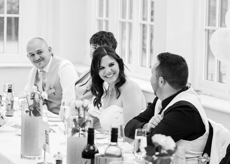A black and white photo of a bride watching her dad give a speech on a wedding day at Swynford Manor in Suffolk Captured by Suffolk Wedding Photographer Hayley Denston Photography