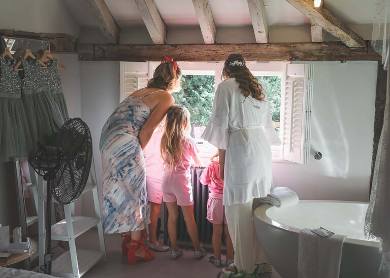 A bride, her sister in law and flowergirls all looking out the window at the wedding set up for her wedding day at Tuddenham Mill in Suffolk Captured by Suffolk Wedding Photographer Hayley Denston Photography