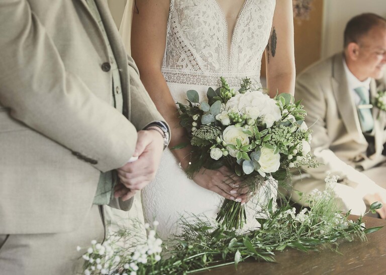 A bride and grooms hands and flowers during a wedding ceremony on a wedding day at Tuddenham Mill in Suffolk Captured by Suffolk Wedding Photographer Hayley Denston Photography