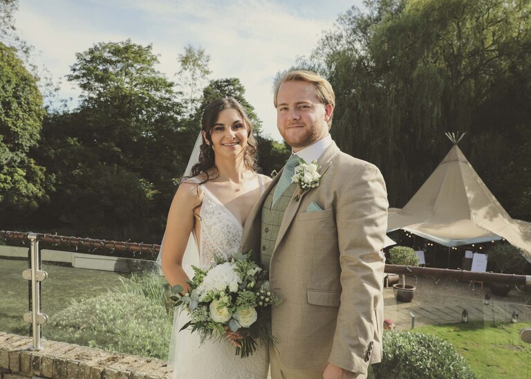 A bride and groom with the mill pond and tipi behind them on their wedding day at Tuddenham Mill in Suffolk Captured by Suffolk Wedding Photographer Hayley Denston Photography