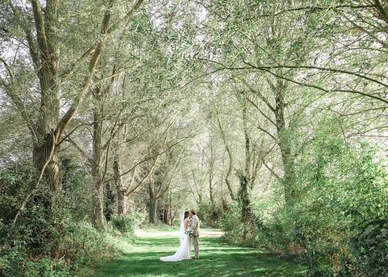A bride and groom kissing in green woodland on their wedding day at Tuddenham Mill in Suffolk Captured by Suffolk Wedding Photographer Hayley Denston Photography