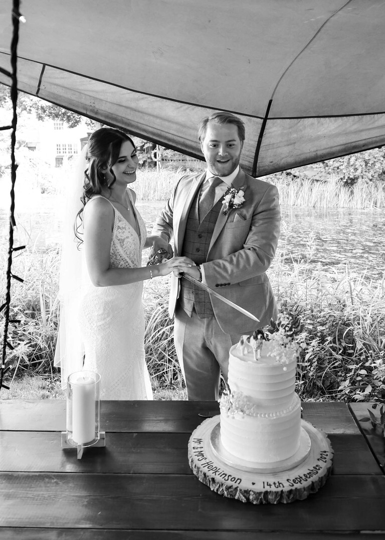 A black and white photo of the bride and groom cutting their wedding cake with a sword on their wedding day at Tuddenham Mill in Suffolk Captured by Suffolk Wedding Photographer Hayley Denston Photography