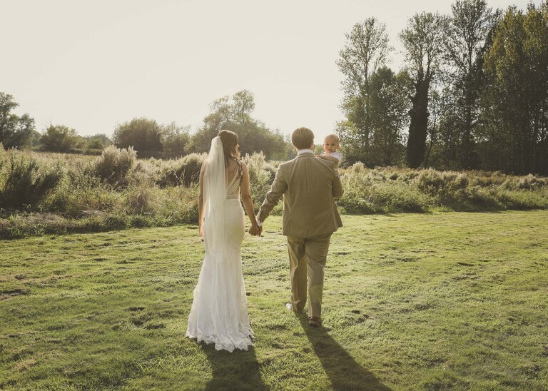 A bride and groom walking into the sunset with their son peeping over the grooms shoulder on their wedding day at Tuddenham Mill in Suffolk Captured by Suffolk Wedding Photographer Hayley Denston Photography
