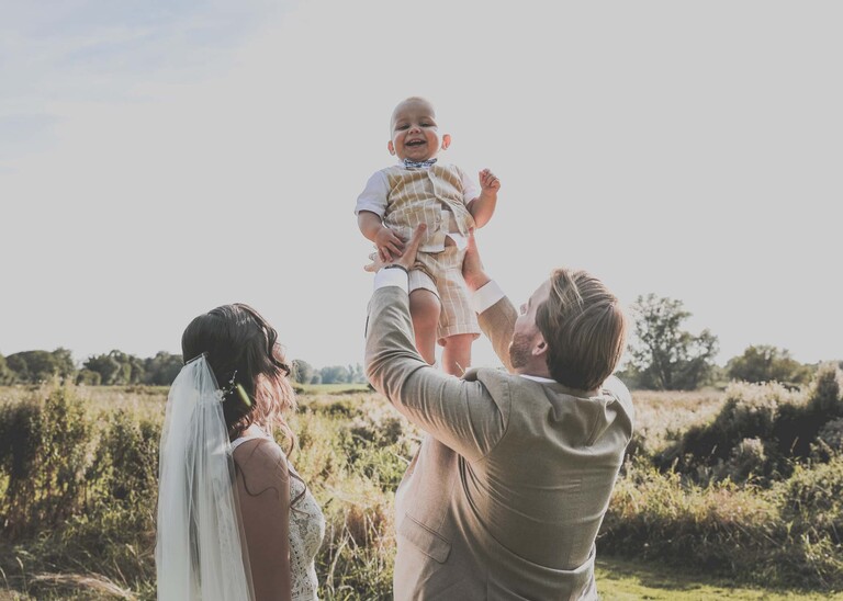 A bride and groom throwing their son into the air on their wedding day at Tuddenham Mill in Suffolk Captured by Suffolk Wedding Photographer Hayley Denston Photography